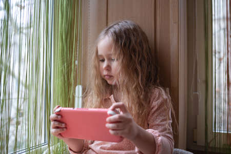 little cute girl plays a game console sitting on the windowsill.