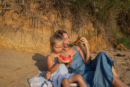 little cute girl with mom eat watermelon sitting on the beach at sunsetの写真素材