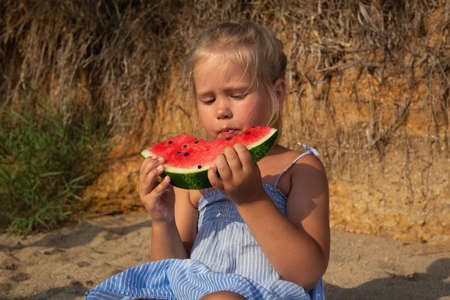 little cute girl eating watermelon sitting on the beach at sunsetの写真素材