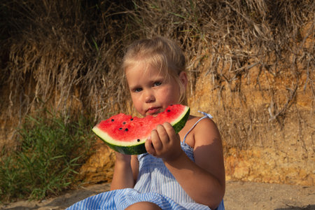 little cute girl eating watermelon sitting on the beach at sunsetの写真素材