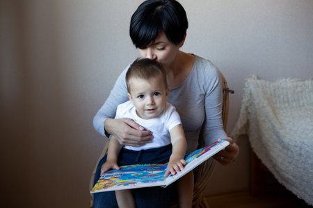 young mom with little son reading book while sitting in a chair in the bedroomの写真素材