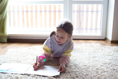 little cute girl draw in an album lying on the floor near a large windowの写真素材