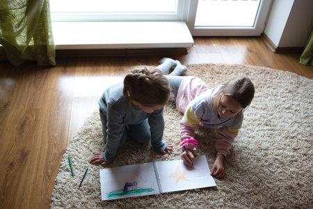 two little cute girls draw in an album lying on the floor near a large windowの写真素材