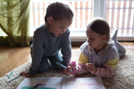 two little cute girls draw in an album lying on the floor near a large windowの写真素材
