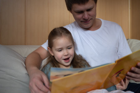 little cute girl with dad read a book sitting in the bedroom on the couchの写真素材