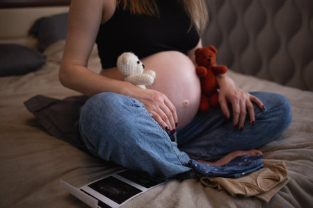 beautiful young pregnant woman sits on the bed in the bedroom and holds her babys toys in her handsの写真素材