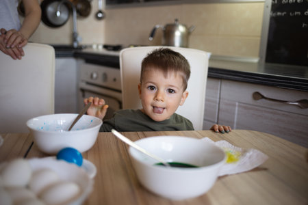 little cute boy paints eggs for easter sitting in the kitchen at the tableの写真素材