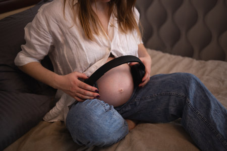 close-up shot. young pregnant woman put headphones on her belly while sitting on the bed in the bedroom.の写真素材