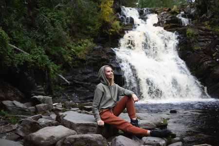 a young beautiful woman tourist sitting against the backdrop of a waterfall surrounded by forestの写真素材