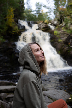 a young beautiful woman tourist sitting against the backdrop of a waterfall surrounded by forestの写真素材