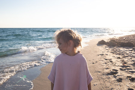 little cute baby girl with tousled hair walks along the sandy beach near the ocean.の写真素材