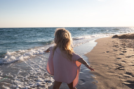 little cute baby girl with tousled hair walks along the sandy beach near the ocean.の写真素材