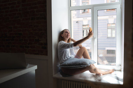 young beautiful woman talking on the phone sitting by the big window in the apartmentの写真素材