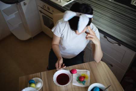 young beautiful woman in bunny ears paints eggs for easter in the kitchenの写真素材