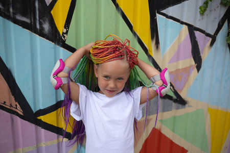 portrait of a little beautiful girl with multi-colored african braids on the background of a colorful wallの写真素材