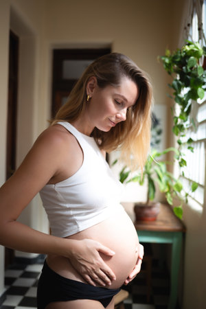 young pregnant woman hugging her big cute tummy while standing by the big window in vintage apartment. the concept of self-love and harmonyの写真素材