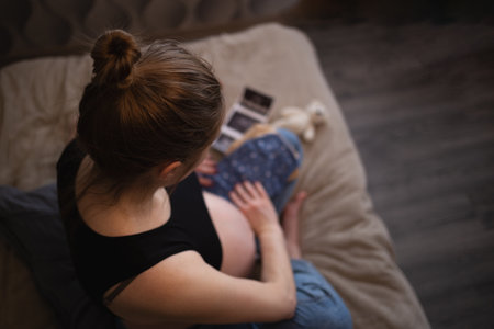 beautiful young pregnant woman sits on the bed in the bedroom and holds her babys tiny clothes in her handsの写真素材