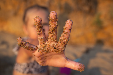 close-up shot. a little girl stretches forward her hand all soiled in the sand, sitting on the seashoreの写真素材