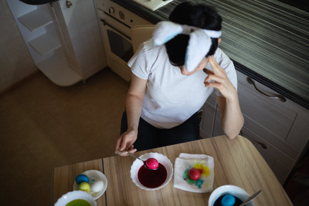 young beautiful woman in bunny ears paints eggs for easter in the kitchenの写真素材