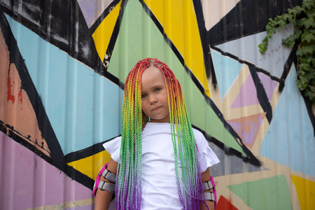 portrait of a little beautiful girl with multi-colored african braids on the background of a colorful wallの写真素材