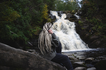 young beautiful Asian woman with African braids against the backdrop of a picturesque waterfall surrounded by forest.の写真素材