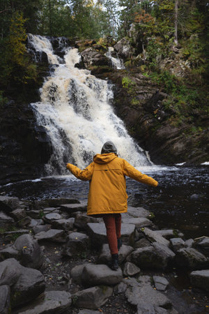 a young beautiful woman tourist stands in a yellow jacket against the backdrop of a waterfall surrounded by forest.の写真素材
