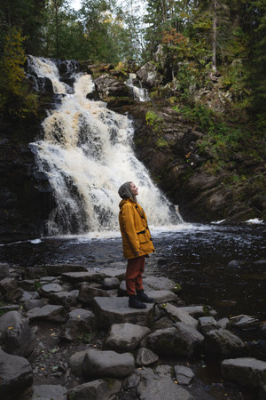 a young beautiful woman tourist stands in a yellow jacket against the backdrop of a waterfall surrounded by forest.の写真素材