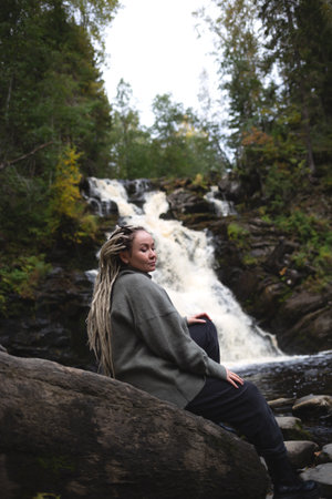 young beautiful Asian woman with African braids against the backdrop of a picturesque waterfall surrounded by forest.の写真素材