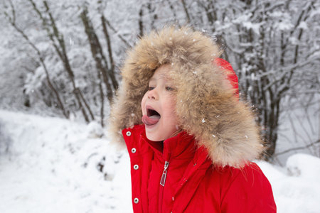 little cute girl in a red jacket catches snowflakes with her mouth in the forest
