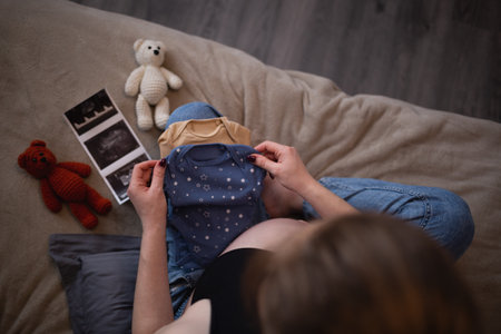 beautiful young pregnant woman sits on the bed in the bedroom and holds her baby's tiny clothes in her hands.の写真素材