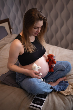 beautiful young pregnant woman sits on the bed in the bedroom and holds her baby's tiny clothes in her hands.の写真素材