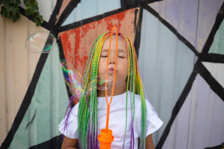 a cute little girl with multi-colored afro pigtails inflates soap bubbles against the background of a beautifully decorated wall.の写真素材