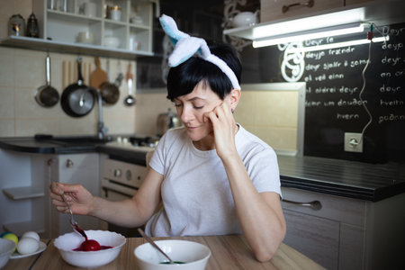 young beautiful woman in bunny ears paints eggs for easter in the kitchenの写真素材