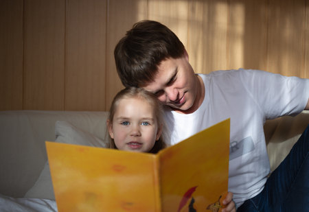little cute girl with dad read a book sitting in the bedroom on the couchの写真素材