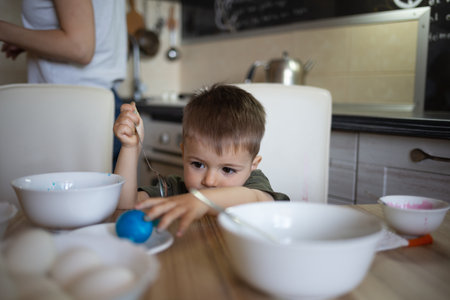 little cute boy paints eggs for easter sitting in the kitchen at the tableの写真素材