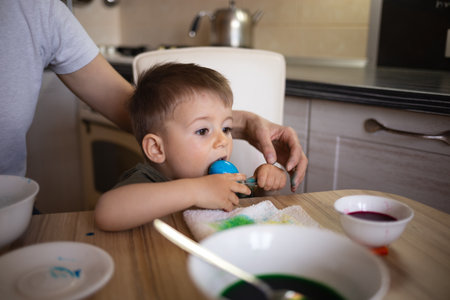 little cute boy paints eggs for easter sitting in the kitchen at the tableの写真素材