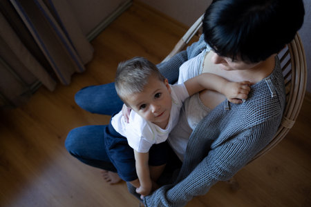 a young woman holds her little son in her arms while sitting in a cozy armchairの写真素材