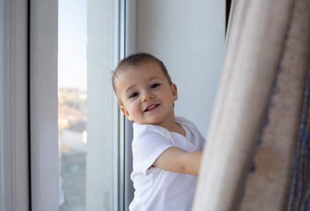 little boy smiling standing on the windowsill by the window behind the curtainの写真素材