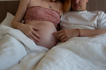 Couple in bed, lovingly touching her pregnant belly, sharing a tender moment of anticipation