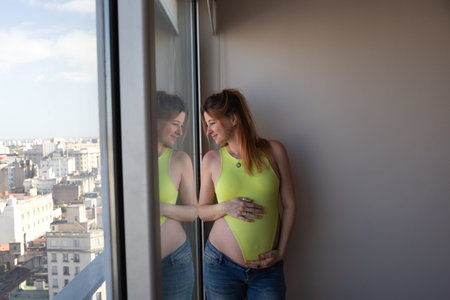 A pregnant woman in a yellow bodysuit smiles, reflecting by the window, anticipating her futureの写真素材
