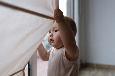 A curious baby pulls the curtain aside, discovering the world beyond the window with wonderの写真素材