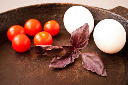 Old cast iron frying pan, cherry tomatoes, eggs and basil leaves. Close upの写真素材
