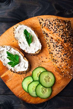 Slices of fresh cucumber, baguette and sandwich with cream cheese and parsley on wooden cutting boardの写真素材