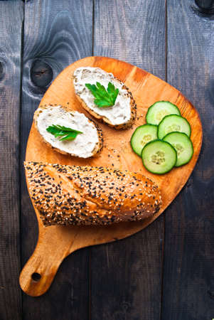 Slices of fresh cucumber, baguette with sesame and sandwich with cream cheese and parsley on wooden cutting boardの写真素材