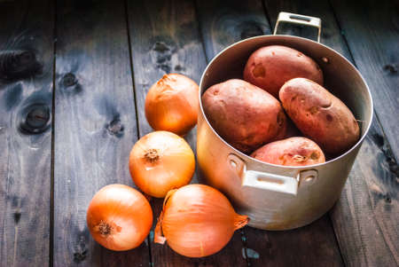 Raw potatoes in a pan and onions beside on a wooden tableの写真素材