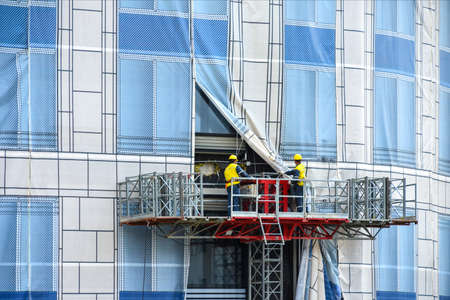 Builders on high-altitude lift work on the wall of a large building covered with colored wallpaperの写真素材