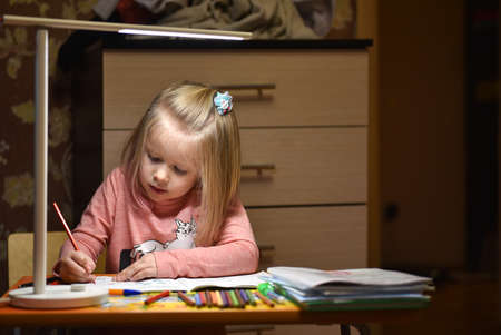 Child preschooler learns to draw and write in notebooks at home in the evening under the light from...
