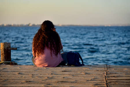 A young girl sits on a sea pier and is sad, view from the back.の写真素材