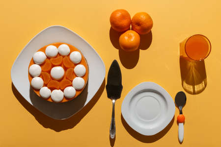 Tangerine cheesecake with meringue discs on white square plate, three tangerines, glass of juice, spatula, dessert plate and spoon on a yellow table illuminated by frontal lightの写真素材