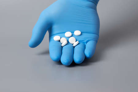 A blue-gloved hand holds pills on a gray background. The doctors blue-gloved hand holds the medicine